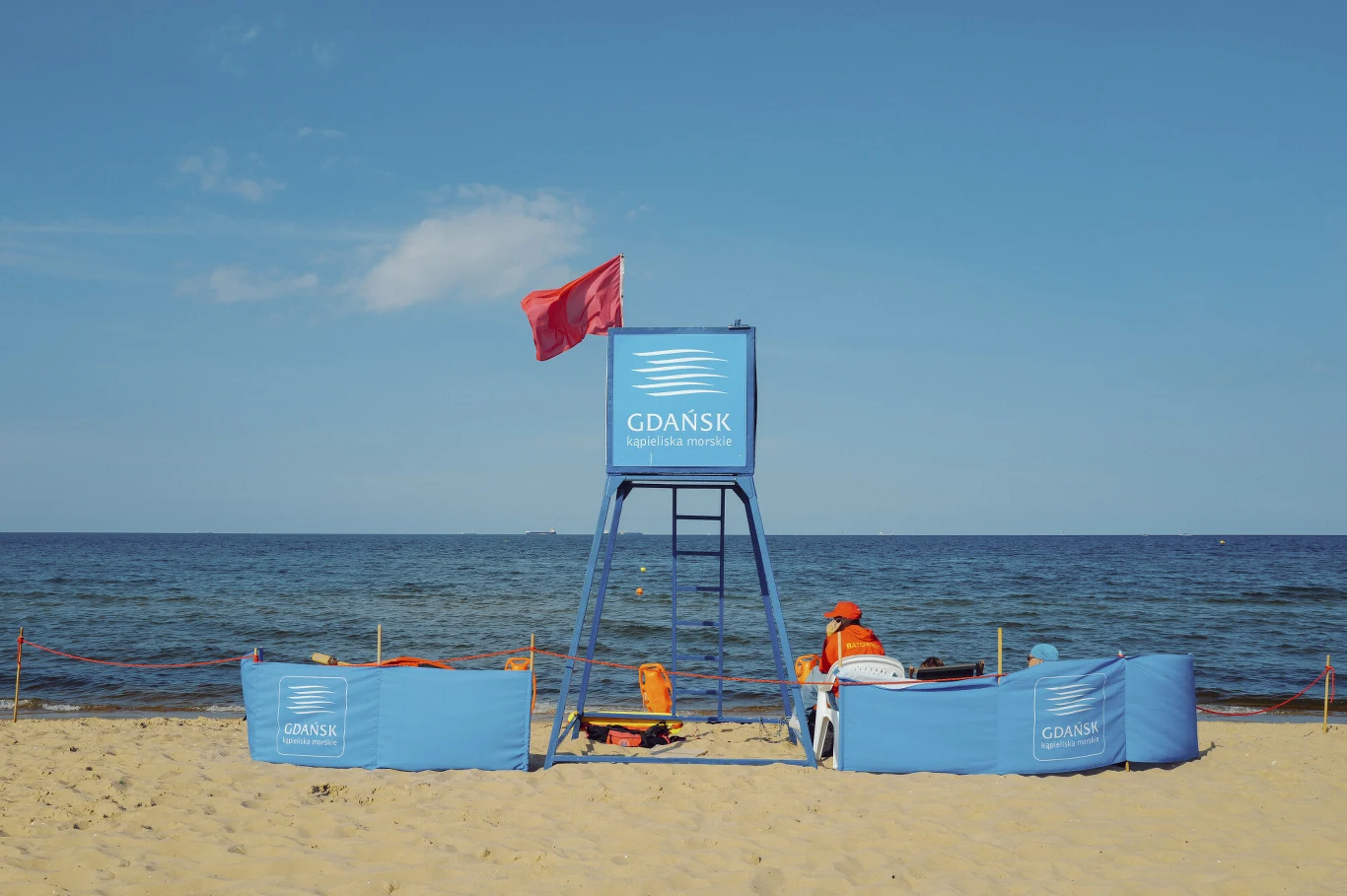 Sinice wróciły nad Bałtyk. Te popularne kąpieliska są zamknięte Czerwona flaga na plaży nad Bałtykiem informująca o zakazie kąpieli z powodu zakwitu sinic. W tle morze i turyści spacerujący po brzegu.
