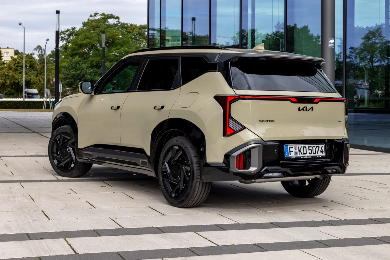 Jasny nowoczesny SUV marki Kia z czarnymi felgami i charakterystycznymi tylnymi światłami LED, zaparkowany na placu przy szklanym biurowcu.