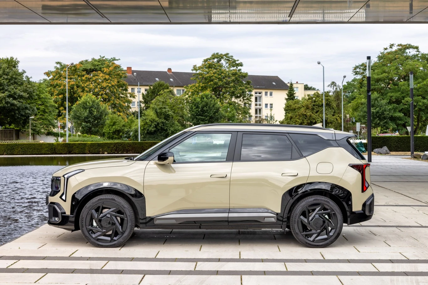 Jasny, nowoczesny SUV z czarnymi detalami i stylowymi felgami stoi zaparkowany na brukowanym placu, w tle miejska zabudowa i zielone drzewa.