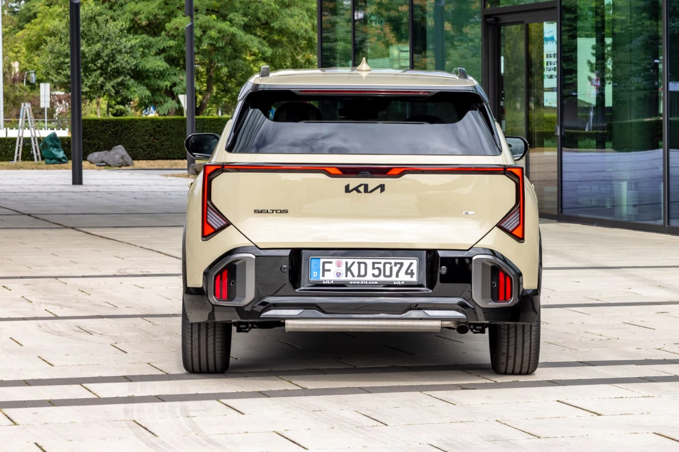 Tył beżowego SUV-a marki Kia z nowoczesnymi, czerwonymi lampami LED oraz niemiecką tablicą rejestracyjną, zaparkowany na betonowej nawierzchni obok szklanej fasady budynku.