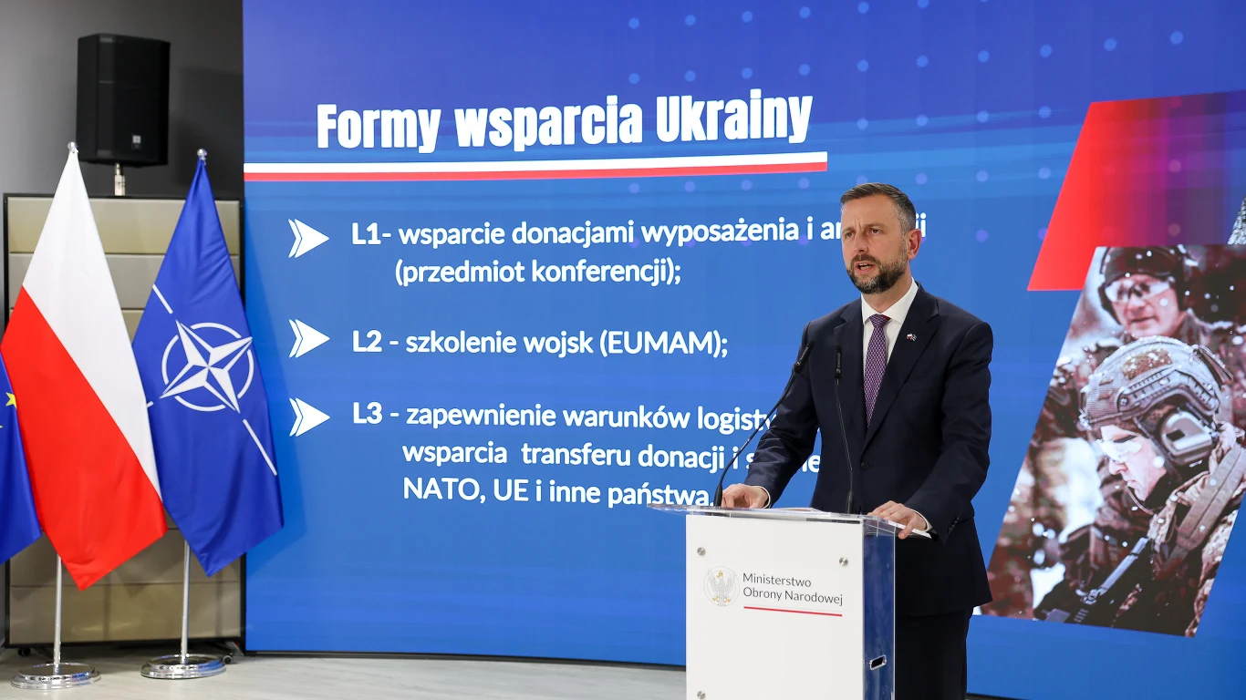 Mężczyzna w garniturze przemawia przy mównicy z logo Ministerstwa Obrony Narodowej, a za nim znajduje się ekran prezentujący formy wsparcia Ukrainy, z flagami Polski i NATO ustawionymi po lewej stronie.