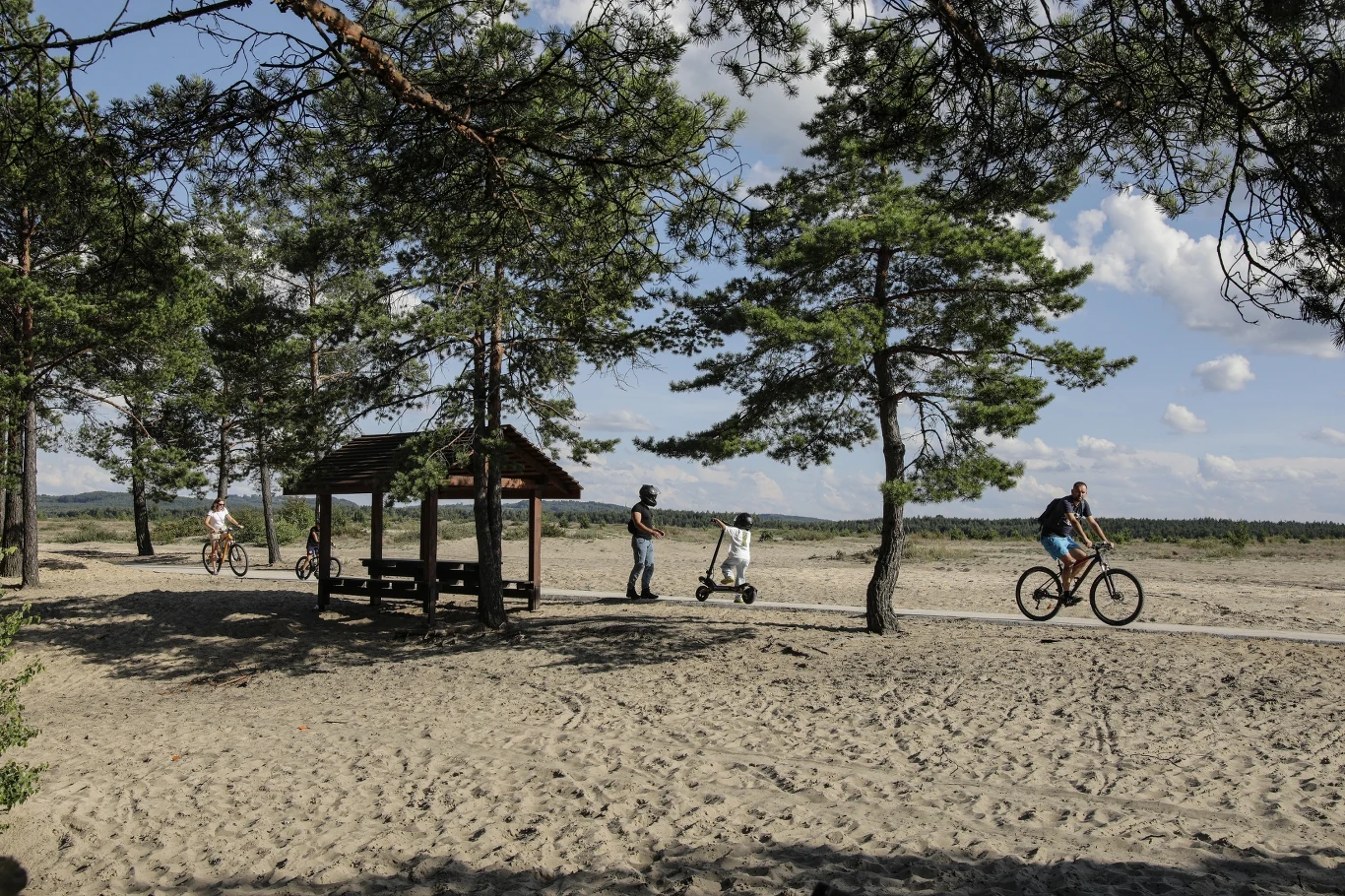 Velo Pustynia jest idealną propozycją dla rodzin z dziećmi i osób poszukujących tras na rekreacyjne przejażdżki. Kilka osób na rowerach i hulajnodze poruszających się ścieżką biegnącą przez piaszczysty teren z rozproszonymi sosnami, obok drewnianej altany.