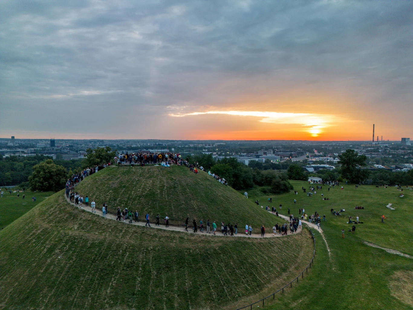 Zielone wzgórze otoczone ścieżką, po której spacerują ludzie, z panoramą miasta i zachodem słońca w tle.