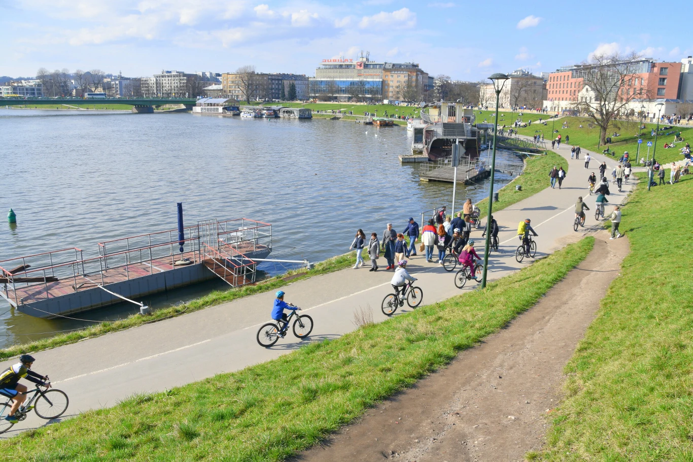 Ludzie spacerują i jeżdżą na rowerach szeroką ścieżką biegnącą wzdłuż brzegu rzeki, otoczeni zieloną trawą i miejską zabudową w tle.