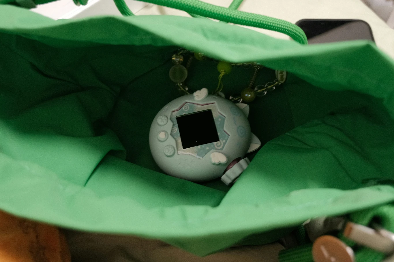 Zielona torebka z elektroniczną zabawką w stylu Tamagotchi na breloczku oraz fragmentem telefonu komórkowego.