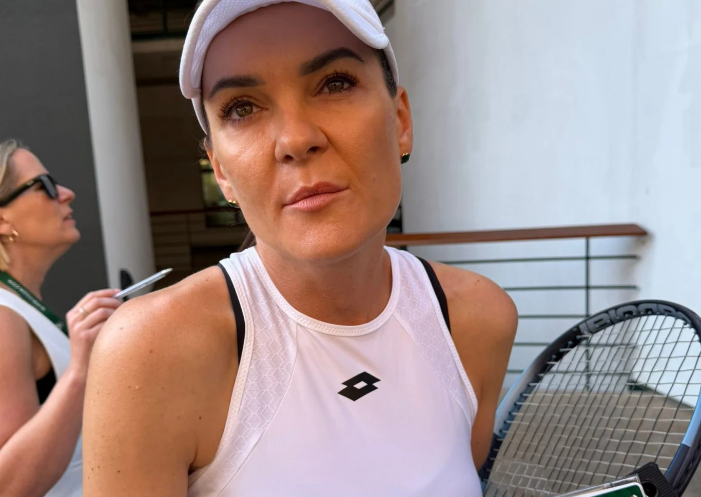 Kobieta w białej czapce z daszkiem i sportowej koszulce stoi z rakietą tenisową, w tle druga osoba z notesem.