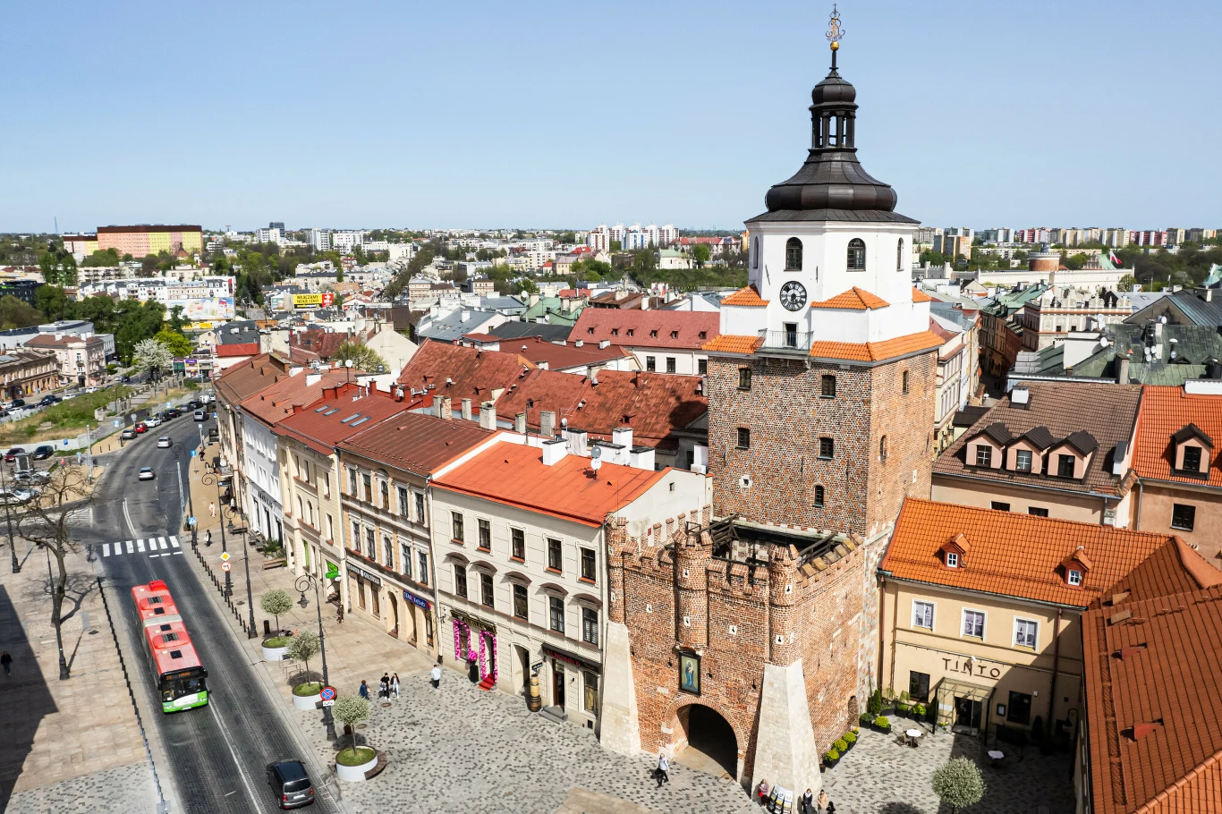 Brama Krakowska z charakterystyczną wieżą, otoczona historycznymi kamienicami z czerwonymi dachami, przy brukowanej ulicy z przejeżdżającymi autobusami i spacerującymi ludźmi.