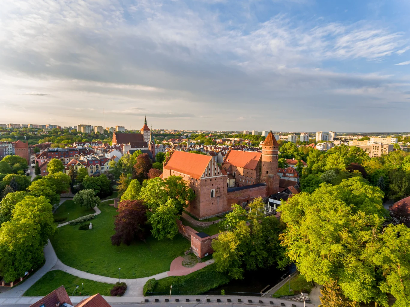 Ceglany zamek otoczony rozległymi zielonymi drzewami, w tle rozpościera się panorama miasta z licznymi budynkami mieszkalnymi i obiektami sakralnymi.