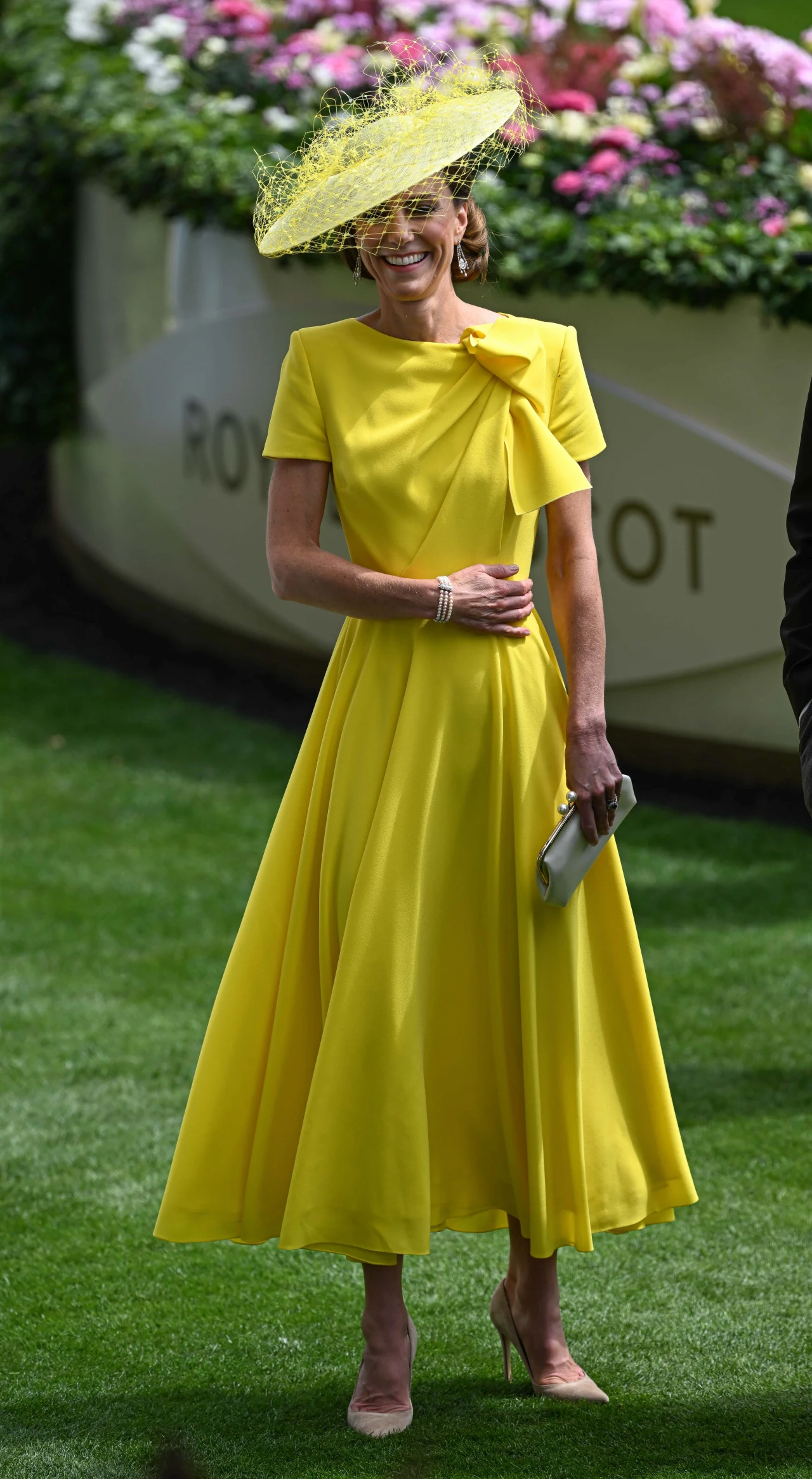 Kobieta w eleganckiej, żółtej sukience do połowy łydki z kokardą na ramieniu, w dopasowanym kapeluszu z siateczką, trzymająca srebrną kopertówkę i pozująca na zielonym trawniku.