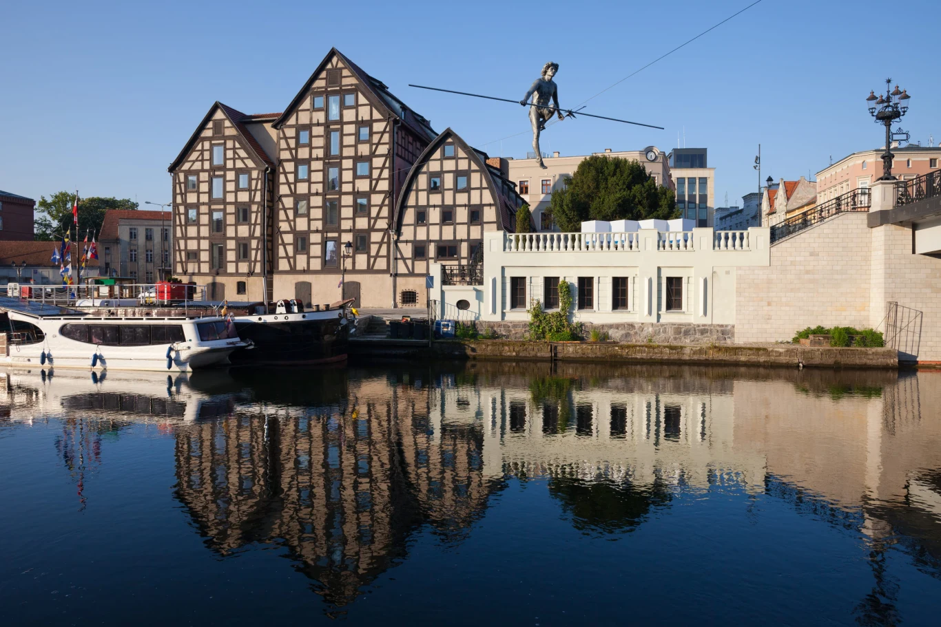 Zabytkowe spichrze z muru pruskiego nad rzeką, obok białego modernistycznego budynku, z rzeźbą akrobaty balansującego na linie nad wodą; na brzegu cumują białe łodzie odbijające się w tafli rzeki.
