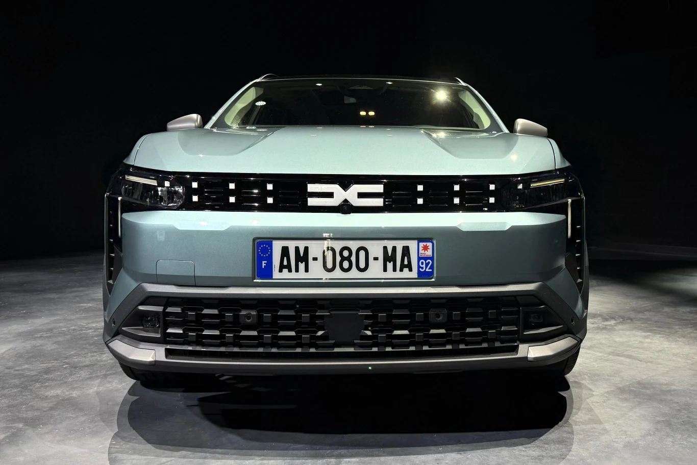 Nowoczesny samochód elektryczny o jasnoniebieskim nadwoziu z wyrazistym, poziomym grillem oraz charakterystycznym logo umieszczonym na środku maski, widoczny z przodu na neutralnym tle.