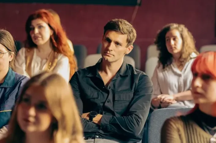 Mężczyzna w ciemnej koszuli siedzący na widowni zwrócony przodem z poważnym wyrazem twarzy, wokół niego inne osoby skupione na słuchaniu.