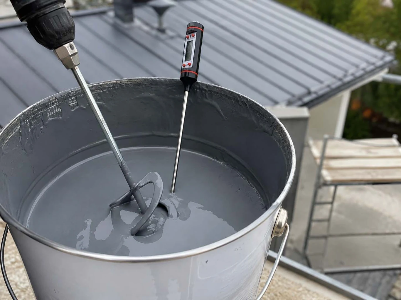 Metalowe wiadro wypełnione szarą farbą lub masą hydroizolacyjną, mieszadło zamontowane na wiertarce oraz termometr zanurzony w płynie, w tle fragment dachu i rusztowania.