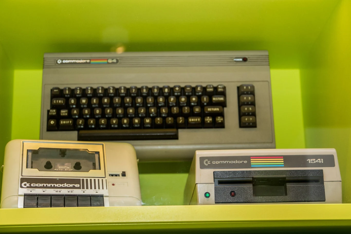 Komputer Commodore 64 z widoczną klawiaturą, magnetofon kasetowy i stacja dyskietek 1541 ustawione obok siebie na zielonym tle.