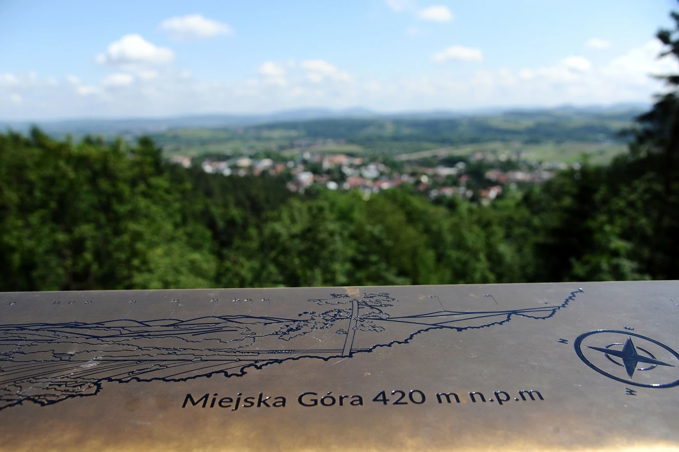 Metalowa tablica informacyjna z napisem 'Miejska Góra 420 m n.p.m', w tle rozległa panorama miasta i zielonych wzgórz porośniętych lasem.