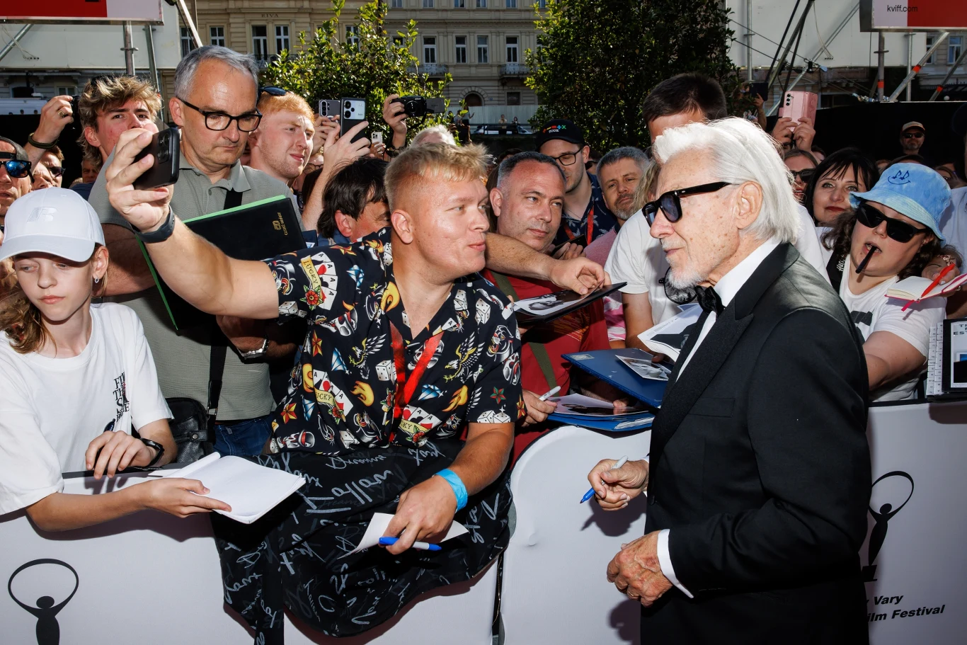 Elegancko ubrany mężczyzna w okularach przeciwsłonecznych pozuje do zdjęcia z fanami, którzy otaczają go z telefonami i notesami, prosząc o autografy na czerwonym dywanie podczas wydarzenia plenerowego.
