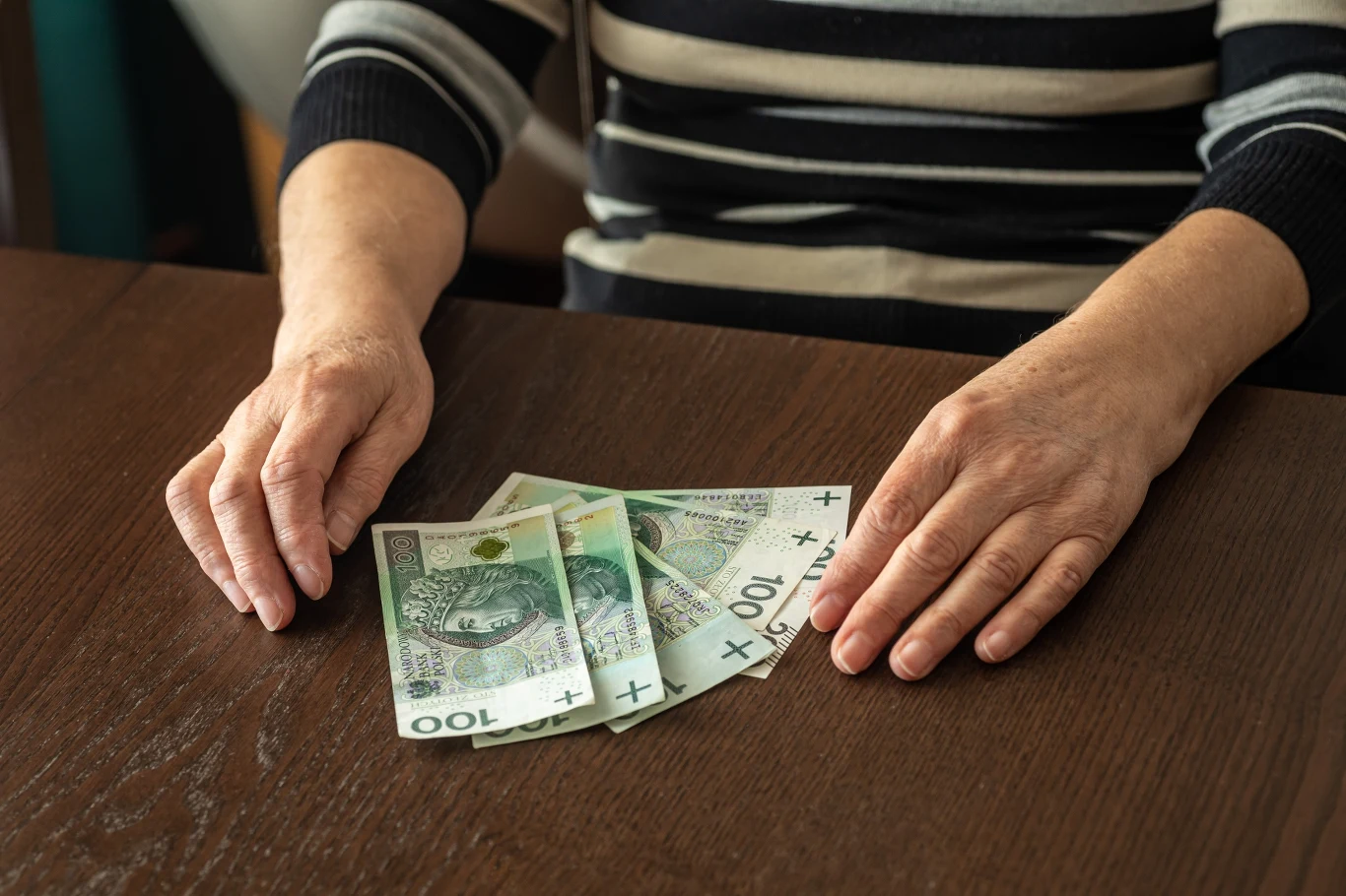 Wielu seniorów nie składa wniosku, choć 500 plus im się należy Ręce osoby w średnim wieku opierające się na stole obok czterech banknotów 100 złotych.