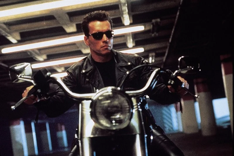 Arnold Schwarzenegger w scenie z filmu "Terminator 2: Dzień sądu" Arnold Schwarzenegger w czarnej kurtce i okularach prowadzi motocykl w industrialnym otoczeniu w filmie "Terminator 2".