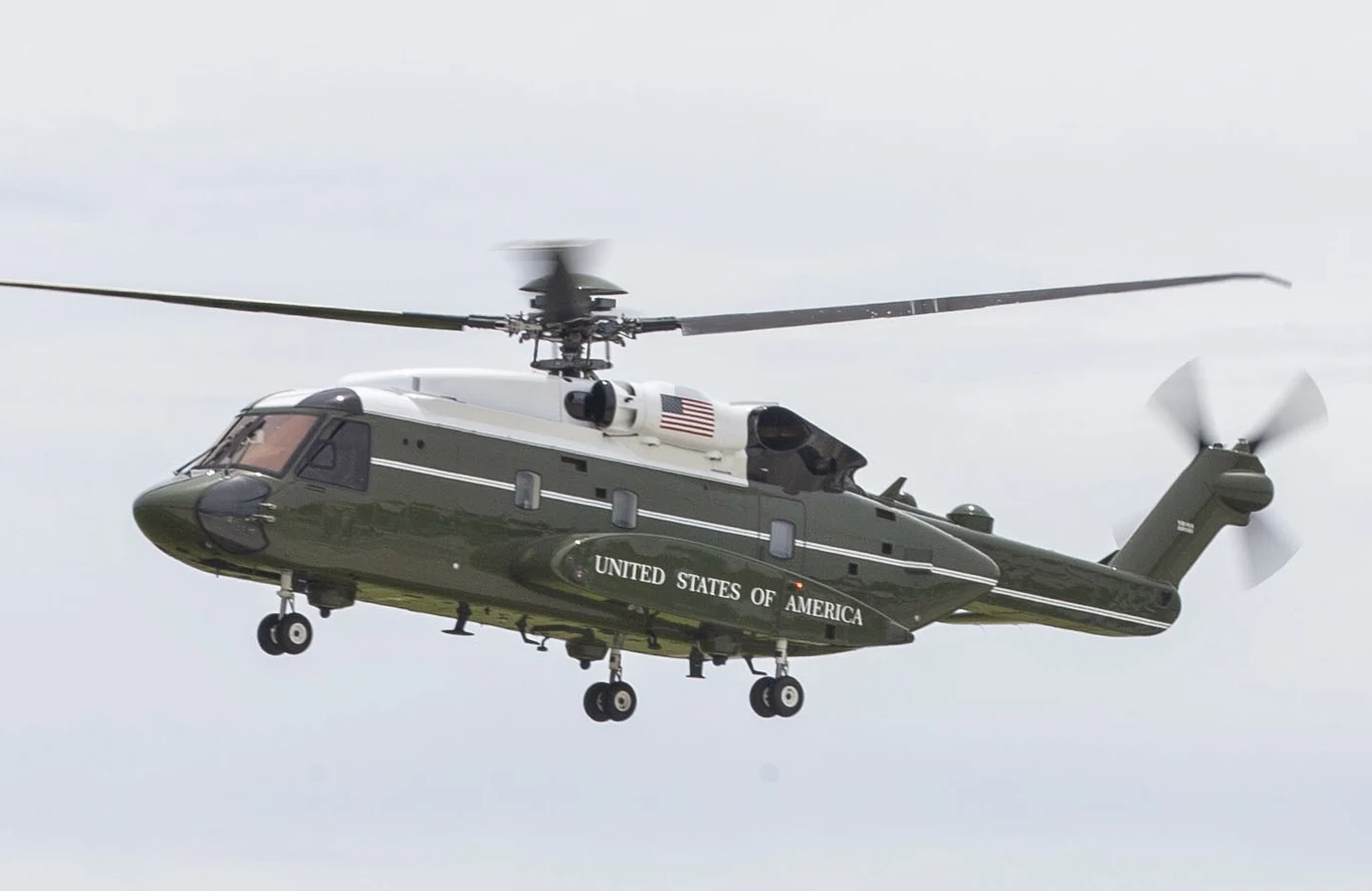 Wojskowy helikopter z charakterystycznym malowaniem, flagą Stanów Zjednoczonych i napisem 'United States of America' w locie na tle jasnego nieba.