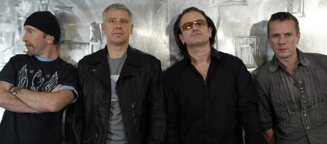 U2 Zespół U2, czterech mężczyzn w ciemnych ubraniach przy metalicznej ścianie, jeden z nich w okularach przeciwsłonecznych