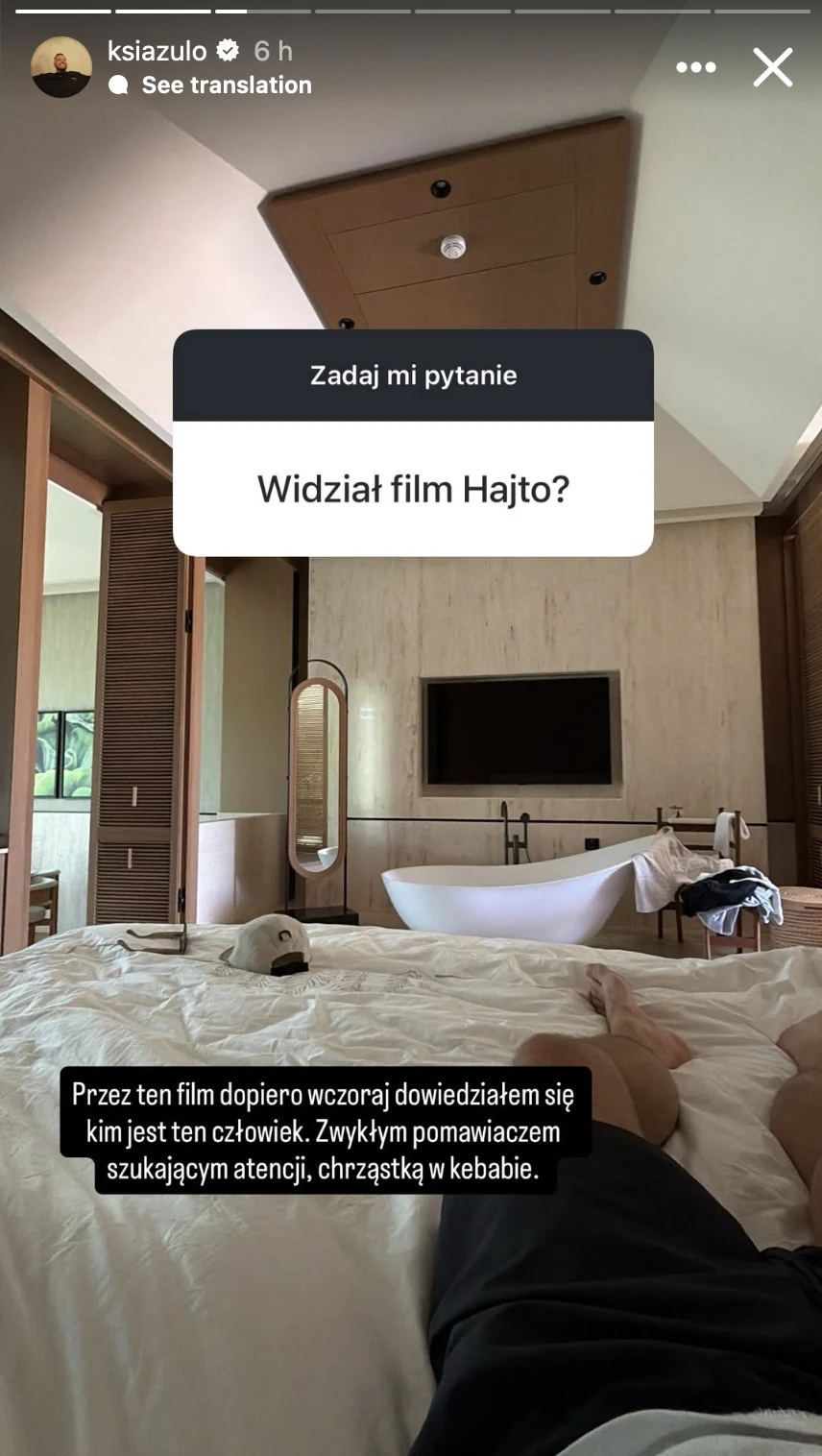 Nowoczesna, przestronna sypialnia z dużym łóżkiem na pierwszym planie, telewizorem zawieszonym na ścianie naprzeciwko oraz dużym lustrem po lewej stronie.