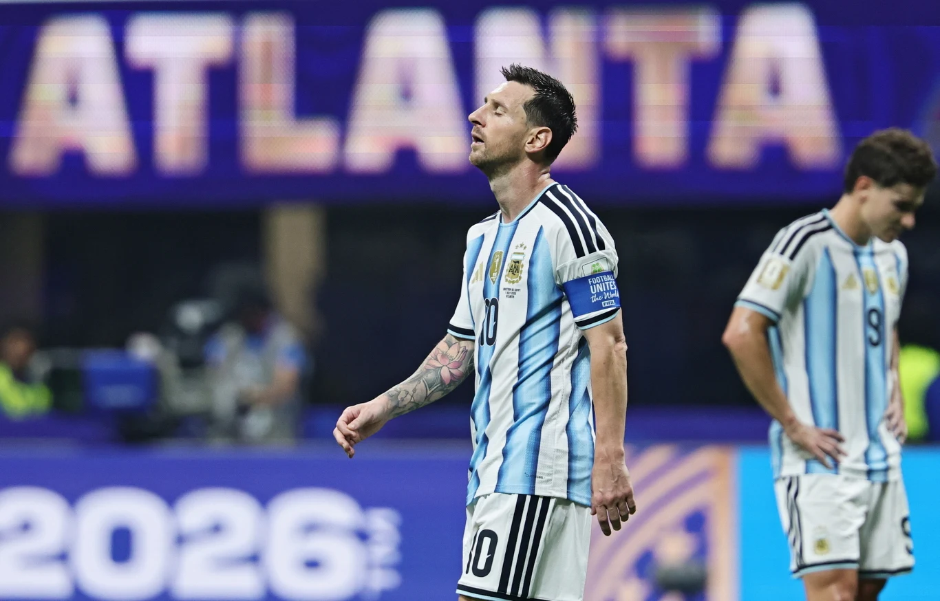 Piłkarz w stroju reprezentacji Argentyny z opaską kapitańską na ramieniu stoi na murawie stadionu, sprawiając wrażenie zamyślonego; w tle duży napis ATLANTA.