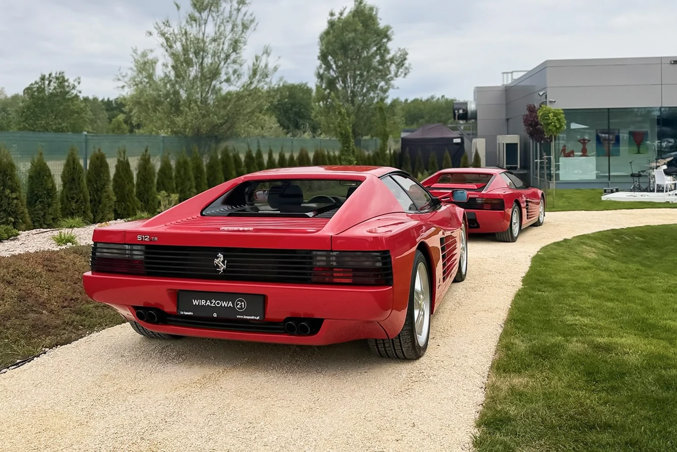 Dwa czerwone samochody Ferrari Testarossa zaparkowane na jasnej żwirowej alejce przy nowoczesnym budynku otoczonym zielenią.