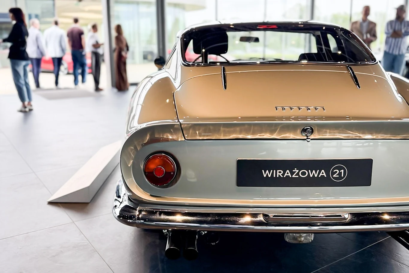 Zabytkowy samochód sportowy Ferrari w kolorze srebrnym zaparkowany w nowoczesnym wnętrzu, w tle kilka osób rozmawia w przestronnej, jasnej sali.