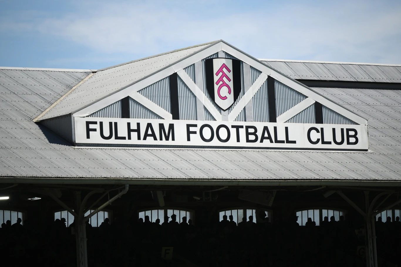 Biała fasada stadionu z dużym szyldem Fulham Football Club pod trójkątnym dachem oraz herbem klubu na górze konstrukcji.