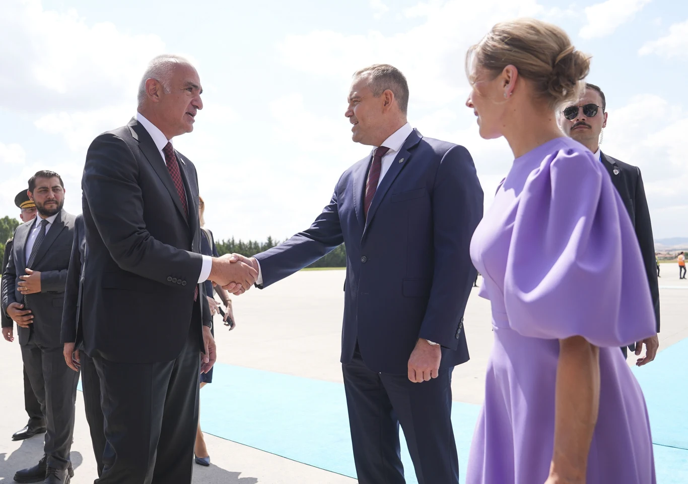 Bufiaste rękawy to romantyczny, a zarazem niezwykle elegancki akcent stylizacji Dwóch elegancko ubranych mężczyzn wymienia uścisk dłoni na płycie lotniska, obok stoi kobieta w fioletowej sukience, a w tle widoczni są inni uczestnicy oficjalnego spotkania.