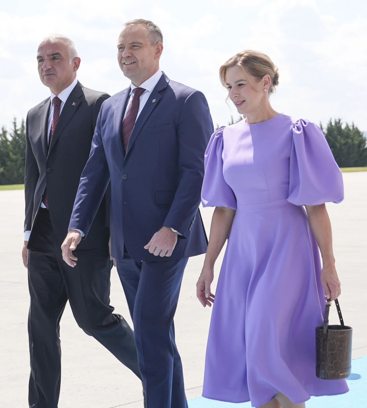 Sukienka w kolorze lawendowym doskonale sprawdzi się tego lata Trzech elegancko ubranych dorosłych idzie razem po wyłożonym niebieskim dywanie, kobieta ma fioletową sukienkę z bufiastymi rękawami i trzyma torebkę, mężczyźni noszą ciemne garnitury i krawaty.