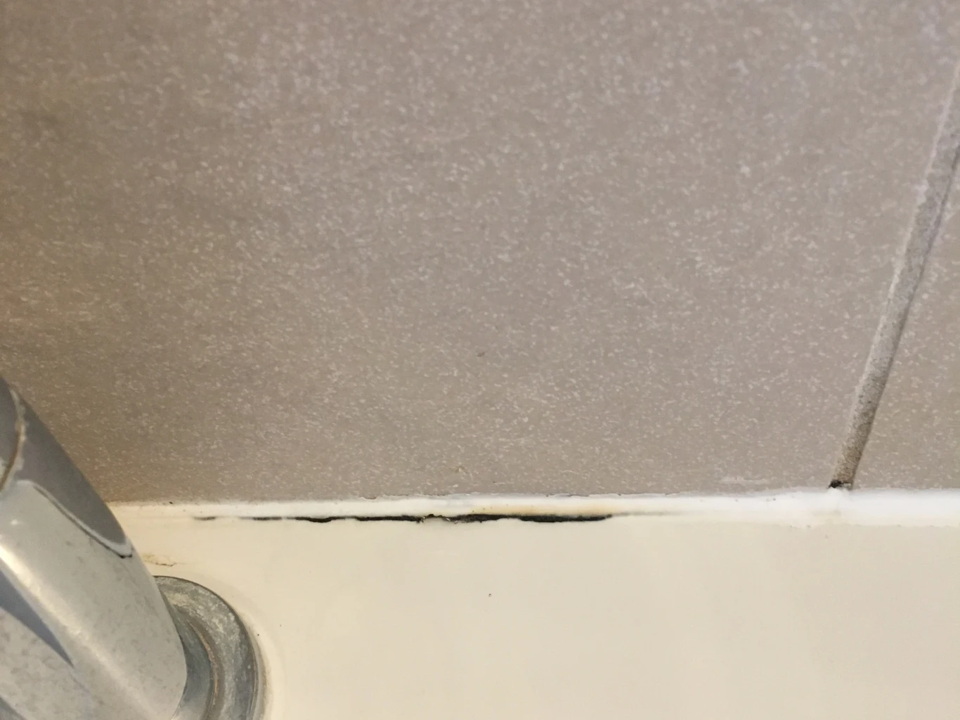 Zabrudzony silikon między białą wanną a szarą płytką ceramiczną, widoczny niewielki zaciek pleśni w spoinie.