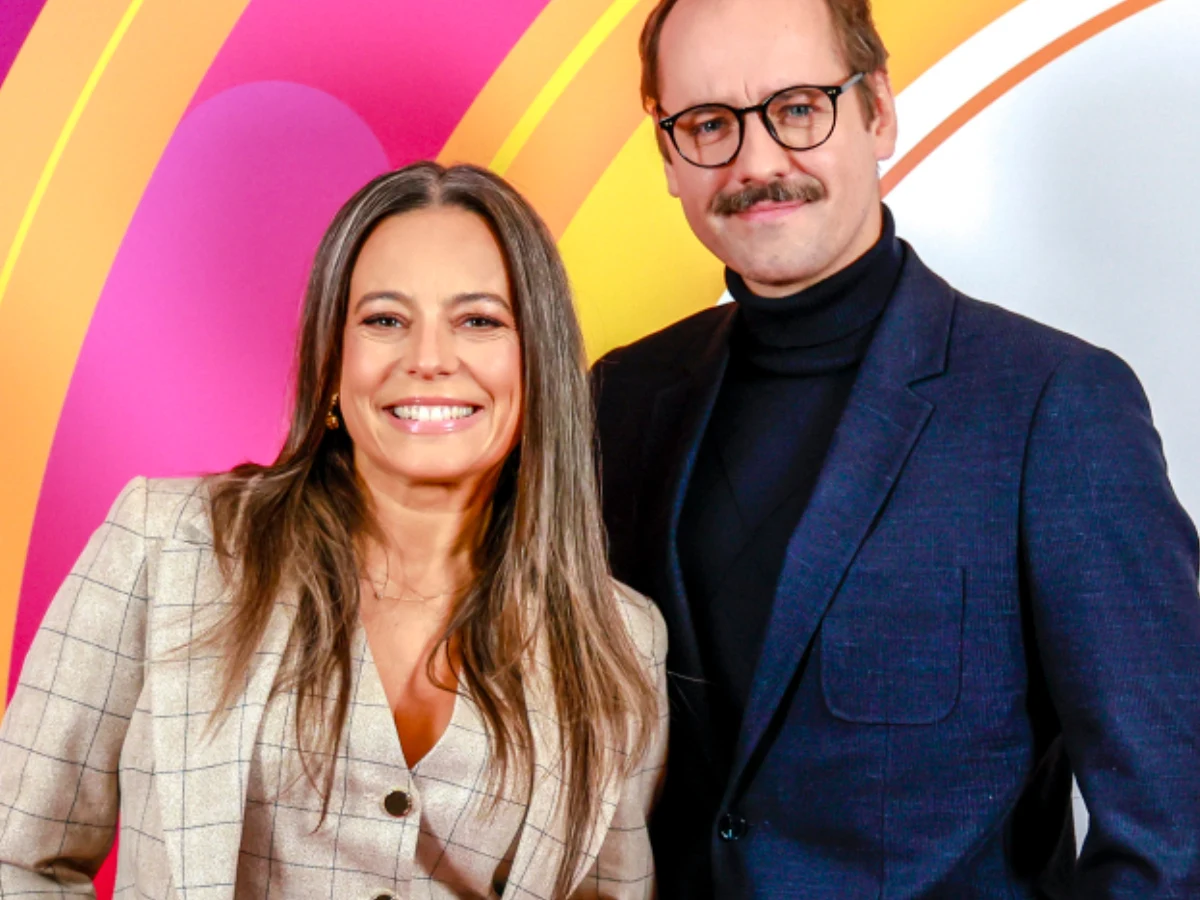 Anna Mucha i Jakub Wons Anna Mucha w marynarce w kratę i Jakub Wons w garniturze stoją razem na tle kolorowych wzorów.