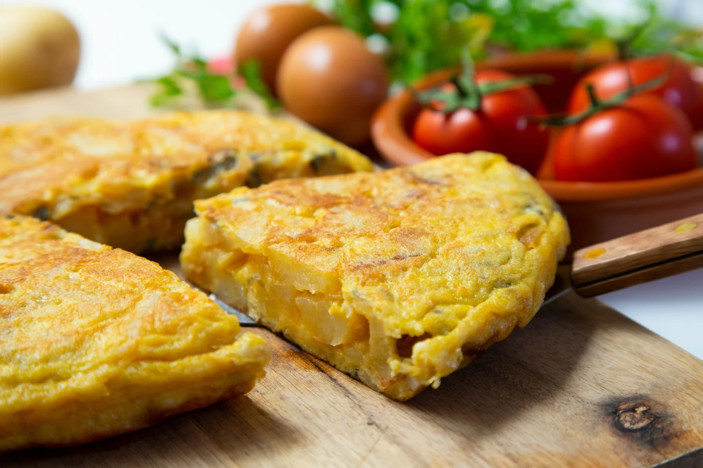 Jak zrobić popularny omlet hiszpański? Pokrojona na porcje hiszpańska tortilla de patatas, obok leżą świeże jajka i pomidory na gałązce.