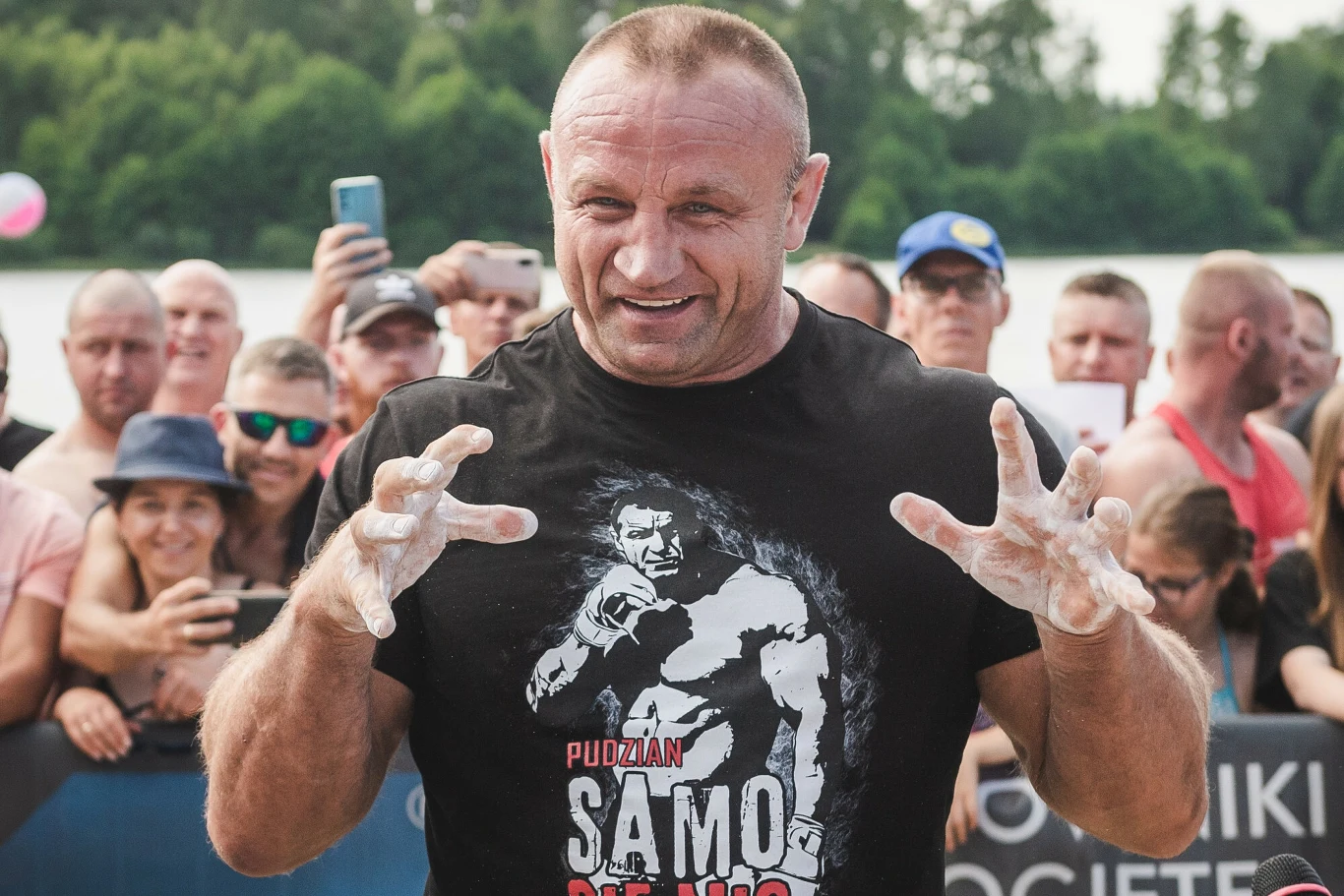 Mariusz Pudzianowski Mariusz Pudzianowski w czarnej koszulce z białym talkiem na rękach, stoi przed grupą ludzi na wydarzeniu plenerowym.