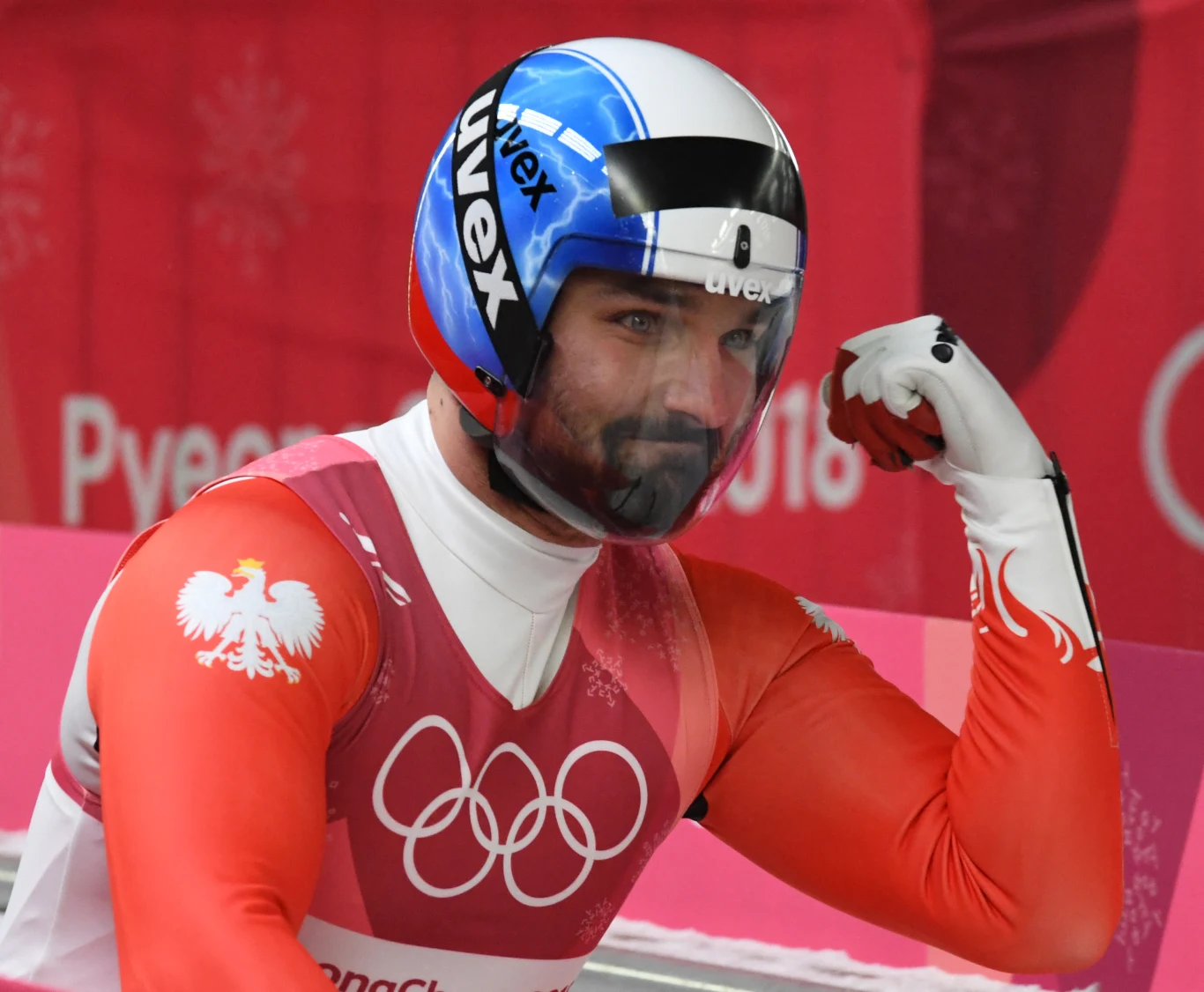 Maciej Kurowski w czasie zimowych igrzysk w Pjongczangu (2018) Sportowiec w kasku i stroju z biało-czerwonym motywem oraz godłem Polski wykonuje gest triumfu, zaciśniętą pięścią prezentując muskuł, na tle logo olimpijskiego.