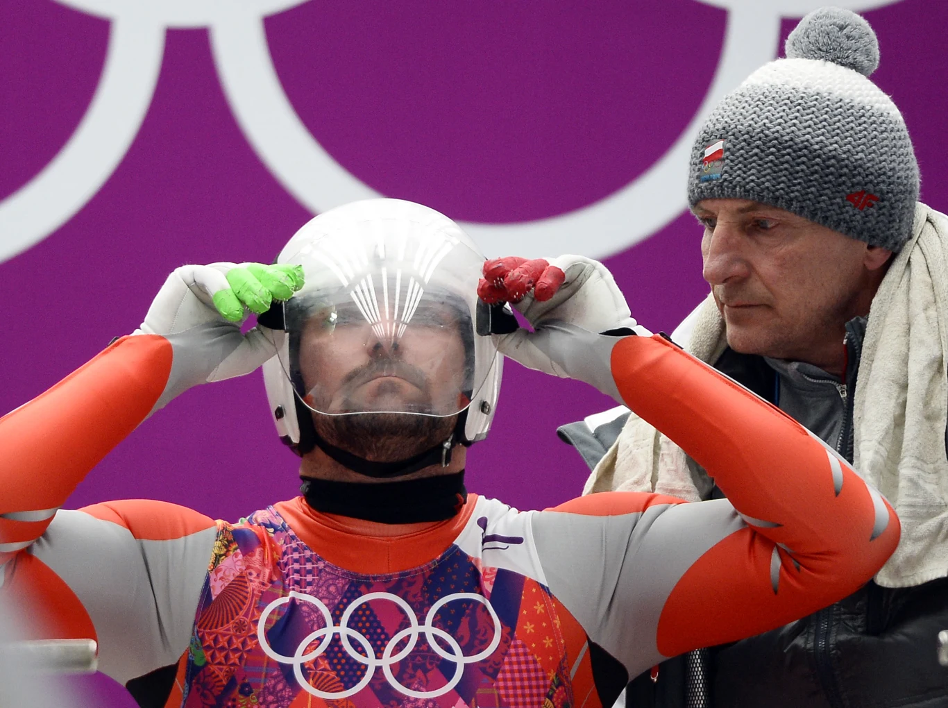 Maciej Kurowski z trenerem Markiem Skowrońskim w czasie zimowych igrzysk w Soczi (2014) Zawodnik w sportowym kombinezonie zakłada kask, obok stoi trener w czapce z orzełkiem i szalikiem na ramieniu.