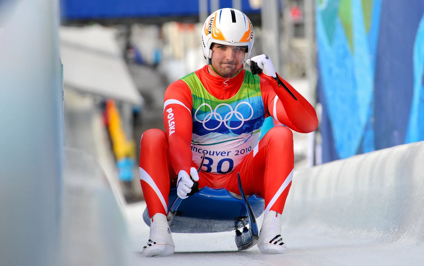 Maciej Kurowski w debiucie olimpijskim w Vancouver (2010) Sportowiec w czerwonym kombinezonie i kasku startuje na sankach na torze lodowym podczas zimowych igrzysk olimpijskich w Vancouver.