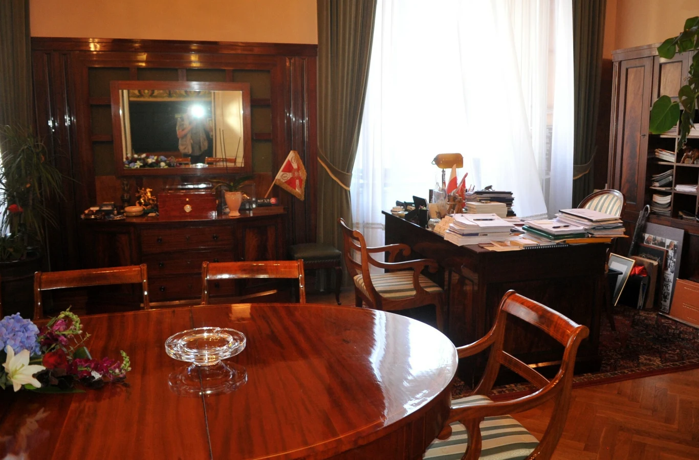 Elegancki gabinet z dużym, drewnianym stołem konferencyjnym na pierwszym planie, obok biurko z lampą, dokumentami i flagami, a w tle regał z segregatorami oraz wysokie okno z białymi zasłonami.