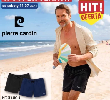 Mężczyzna w czarnych szortach plażowych marki Pierre Cardin, trzymający rakietkę do gry nadmorskiej, spaceruje po piaszczystej plaży przy brzegu morza.