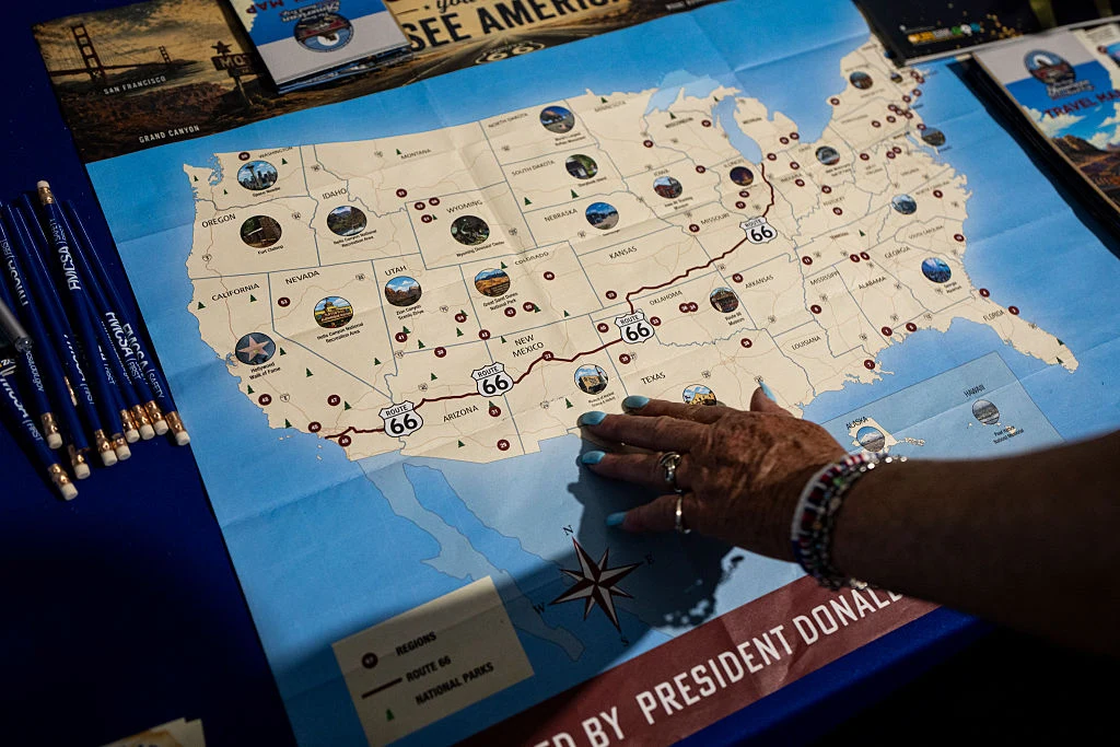 Mapa Stanów Zjednoczonych przedstawiająca trasę Route 66 z zaznaczonymi najważniejszymi punktami na szlaku, ręka wskazująca obszar południowo-zachodni i widoczne wokół mapy gadżety tematyczne, takie jak ołówki oraz broszury.