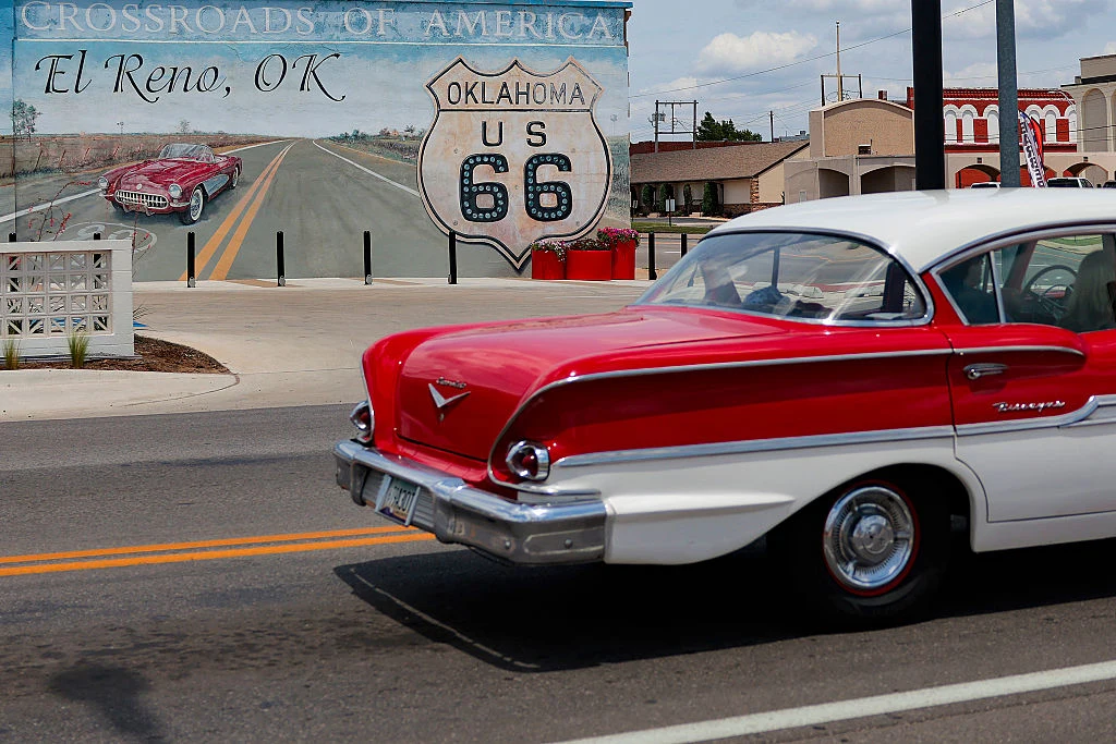 Droga 66 w miejscowości El Reno w Oklahomie Klasyczny czerwono-biały samochód przejeżdża ulicą obok kolorowego muralu z motywem słynnej drogi Route 66 i napisem 'Oklahoma US 66'.