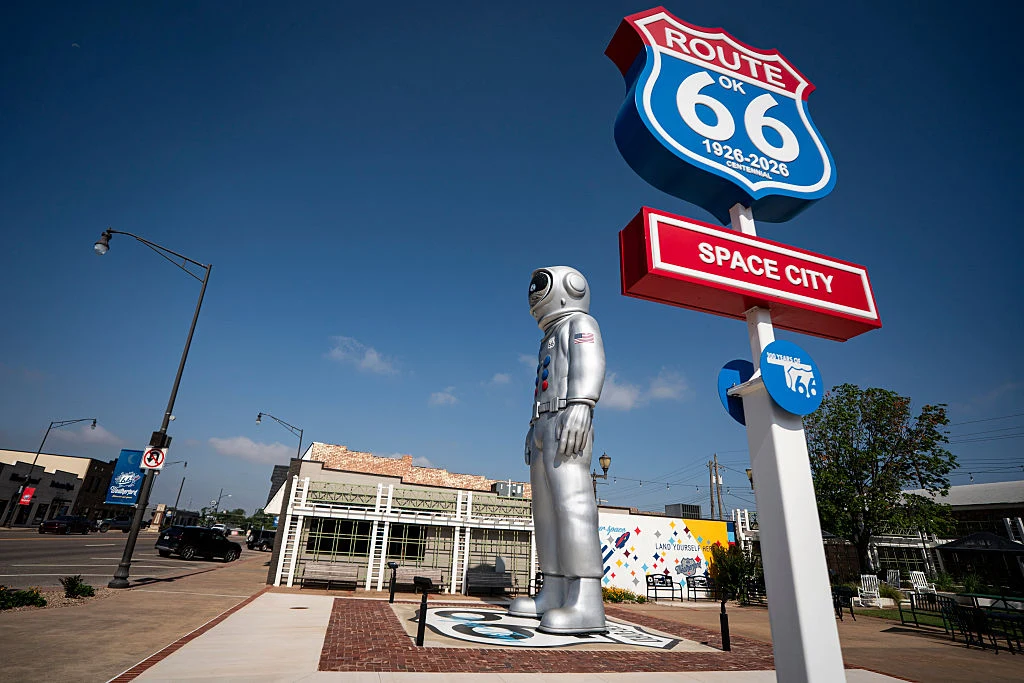 Przy Drodze 66 znajduje się wiele osobliwych atrakcji turystycznych Srebrna rzeźba astronauty w skafandrze kosmicznym stojąca na chodniku obok dużego znaku Route 66 oraz dodatkowymi elementami infrastruktury miejskiej w tle.