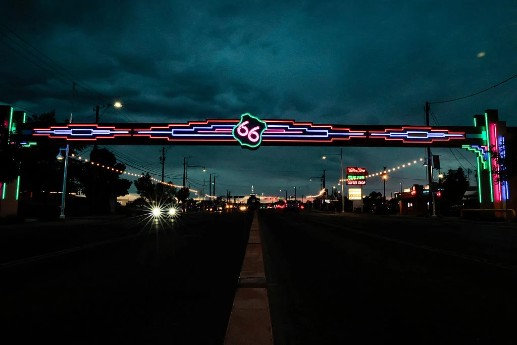 Droga 66 jest jednym z symboli Stanów Zjednoczonych Neonowy łuk z numerem 66 oznaczający słynną trasę Route 66, rozświetlający drogę po zmroku, otoczony ulicznymi światłami i budynkami z neonami w tle.