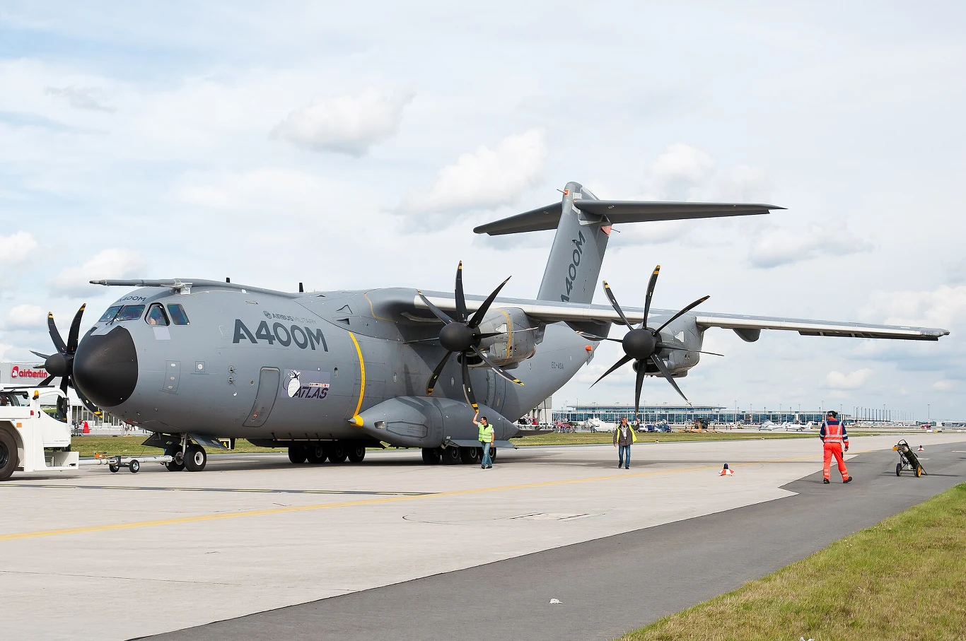 Wojskowy samolot transportowy Airbus A400M Atlas o szarej barwie stoi na płycie lotniska, otoczony przez kilku pracowników w odblaskowych kamizelkach.