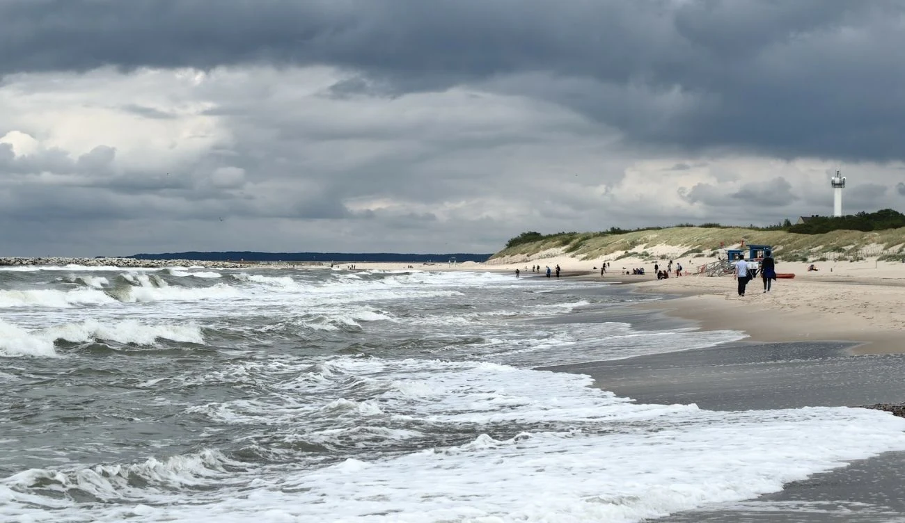 Szeroka plaża nad morzem z falami uderzającymi o brzeg, ludzie spacerujący wzdłuż linii wody, biała latarnia morska widoczna na wydmach pod pochmurnym niebem.