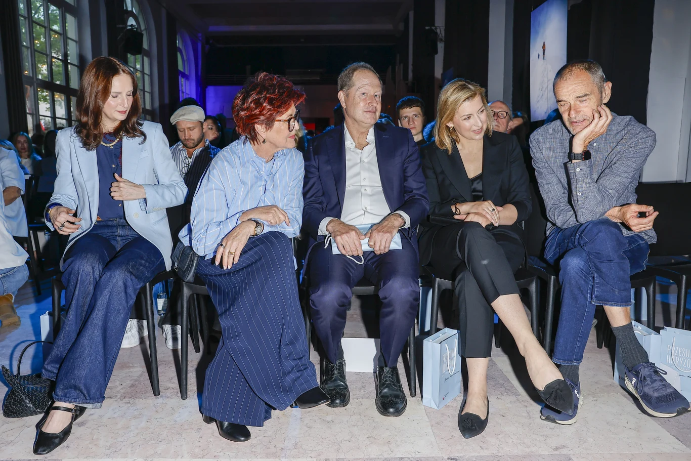 Pięć osób siedzących w pierwszym rzędzie na wydarzeniu, prowadząc ze sobą rozmowy i uśmiechając się, ubrane elegancko, w tle słabo oświetlona sala z innymi uczestnikami.