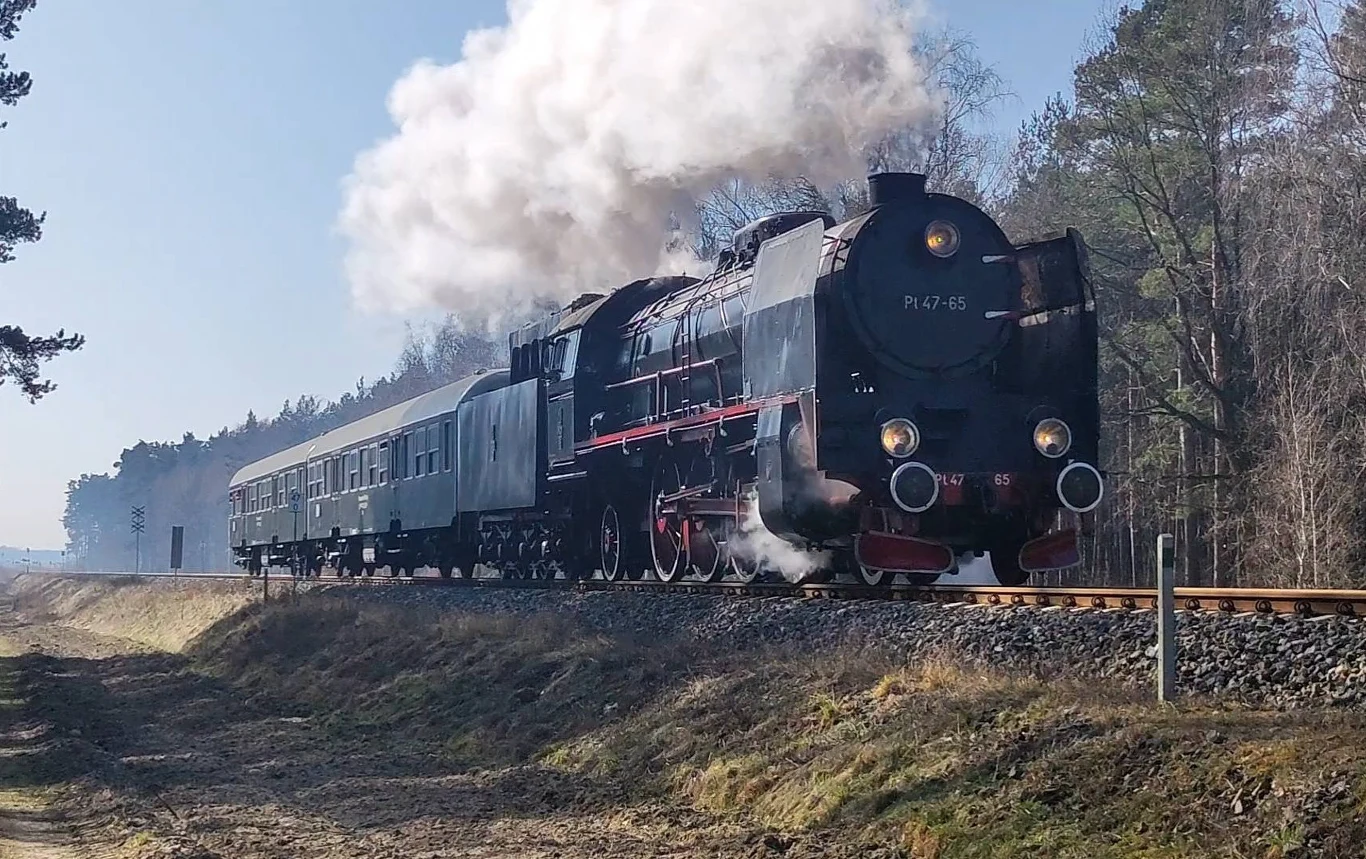 Pociąg retro "Wakacyjny Strażak" odjedzie w weekend. Parowóz Pt47 w akcji. Parowóz Pt47 ciągnący wagon pasażerski po torach, otoczony lasem i polaną, z kłębami białej pary.