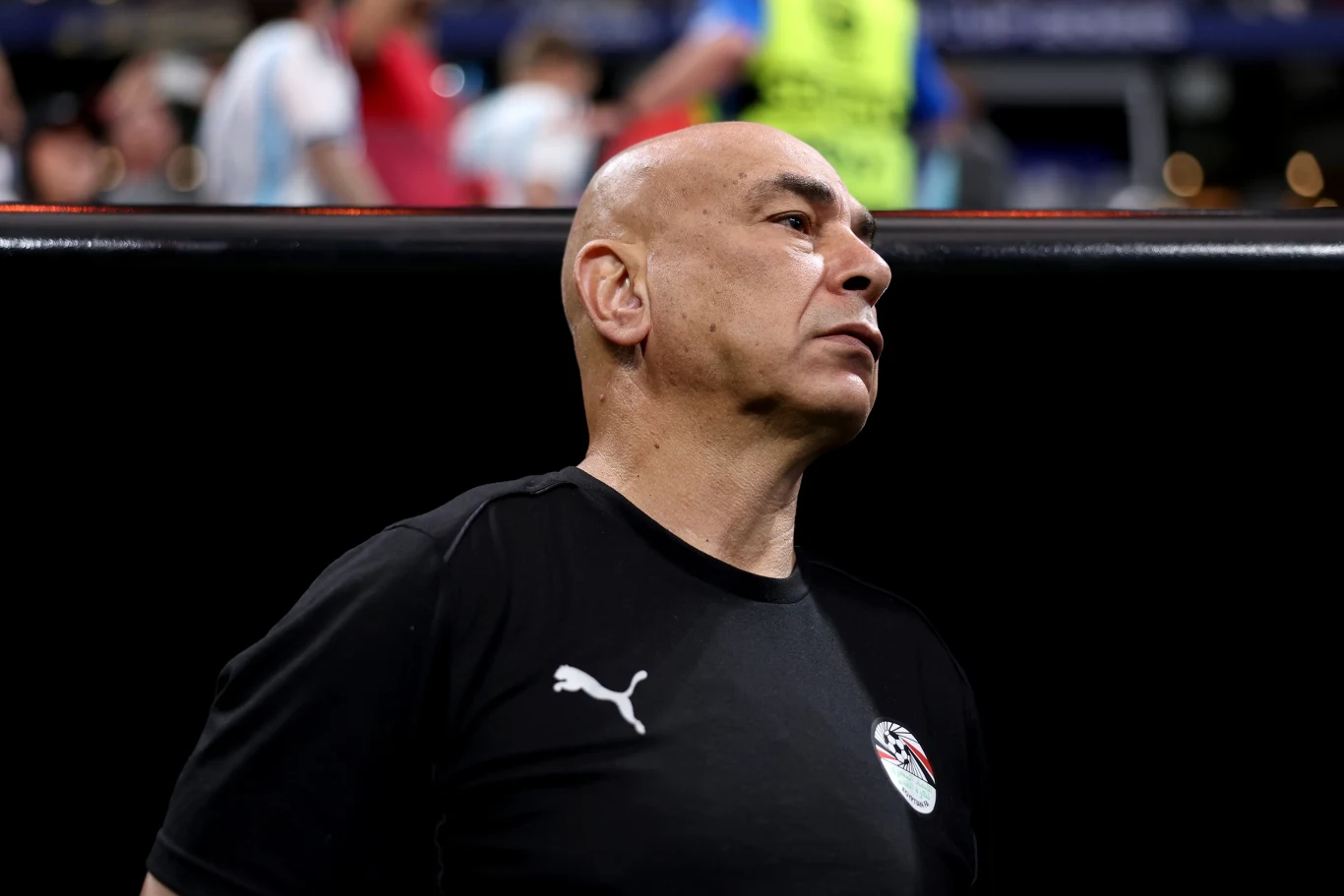 Łysy mężczyzna w czarnej koszulce sportowej z logo Puma oraz godłem Egiptu stoi na tle rozmytego stadionu, patrząc w bok z poważnym wyrazem twarzy.