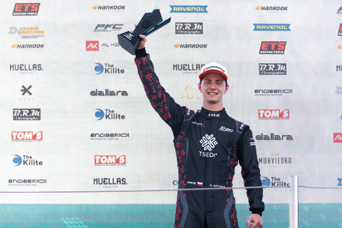 Młody kierowca wyścigowy w czarnym kombinezonie trzyma w górze trofeum, uśmiechając się na podium, w tle ściana z logotypami sponsorów.